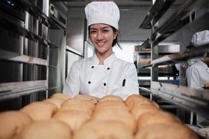 jovem chef feminino asiático em uniforme de cozinheiro branco e chapéu mostrando a bandeja de pão saboroso fresco com sorriso, olhando para a câmera, feliz com seus produtos alimentícios assados, trabalho profissional na cozinha de aço inoxidável. foto