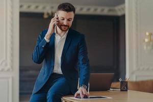 empresário sério falando ao telefone enquanto está sentado na mesa de escritório no escritório moderno foto