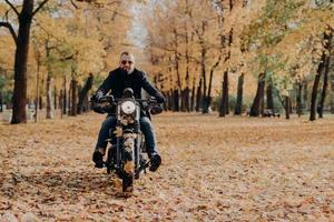 motociclista masculino profissional brutal anda de bicicleta, usa óculos escuros, luvas e jaqueta preta, tem passeio pelo parque outonal, belas paisagens ao fundo com árvores amarelas e folhas caídas ao redor foto