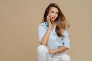 mulher bonita se sente relaxada enquanto se senta na cadeira, vestida com camisa elegante e calça branca, posa sobre cabelos castanhos, copie o espaço para o seu conteúdo promocional. mulher bonita em roupas da moda foto