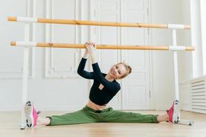 bailarina caucasiana abre as pernas para o lado, mostra aberturas, segura as mãos nas barras, tem aula de balé, usa roupas esportivas e tênis, tem figura perfeita, inclina a cabeça para a direita, estica-se. foto