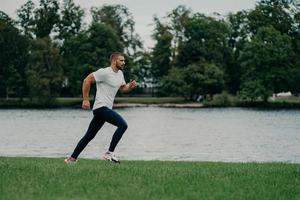 tiro horizontal de homem barbudo ativo bonito vai correndo sendo fotografado em movimento, corre contra o rio e a bela natureza, prepara-se para a maratona. conceito de esporte, bem-estar e treino foto