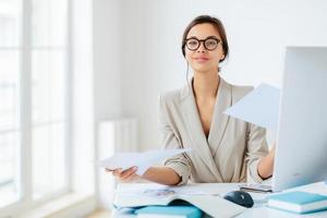 ocupado gerente feminino de sucesso trabalha com papéis no escritório, posa na área de trabalho, usa óculos e roupa formal, ocupado preparando relatório, olha com confiança para a câmera. empresária revisa documentos foto