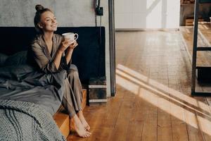 linda garota bonita com uma xícara de café da manhã relaxando enquanto está sentado na cama foto