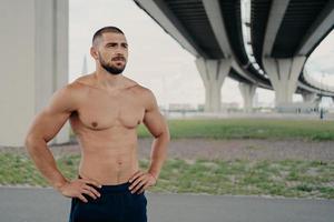 foto de homem barbudo bonito sem camisa com torso nu posa ao ar livre mantém as mãos na cintura concentradas na distância pratica esporte, tem treino matinal, orgulho de ser forte, tem abdominais perfeitos