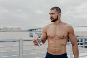 homem barbudo de construção muscular entra para o esporte, segura garrafa de água fresca, focada na distância, posa na ponte perto do rio durante o dia nublado, faz uma pausa após exercícios físicos. treino cardio foto