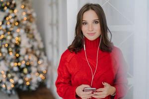 tiro horizontal de jovem mulher caucasiana gosta de música da lista de reprodução, ouve música favorita com fones de ouvido, vestido com suéter vermelho de malha, fica contra o fundo da árvore de natal decorada foto