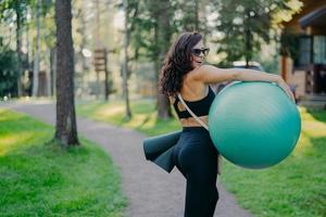 tiro ao ar livre de mulher morena feliz carrega fitball e fitness mat, usa óculos escuros e roupas esportivas, posa no parque verde, estando em boa forma física. estilo de vida saudável e conceito de hobby foto