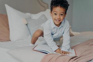 adorável criança afro-americana sorridente na camisa azul clara brincando na cama dos pais foto