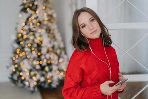 modelo feminino muito jovem ouve música agradável em fones de ouvido, segura celular, vestido com suéter de malha vermelha, posa na sala de estar perto da árvore de natal com espaço em branco para o seu conteúdo publicitário foto