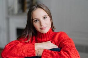 mulher caucasiana com maquiagem, cabelo escuro, vestida de swater vermelho quente, olha diretamente para a câmera com expressão calma, posa no interior contra um fundo desfocado. namorada relaxada gosta de tempo livre foto