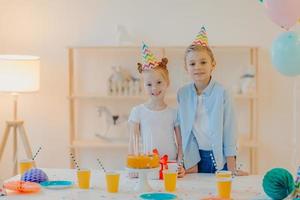 menino feliz e sua pequena irmã ruiva vestidos com roupas festivas, chapéus de festa, comemoram o aniversário juntos, cercados de bolo, presente e copos de bebida, têm bom humor durante ocasiões especiais foto