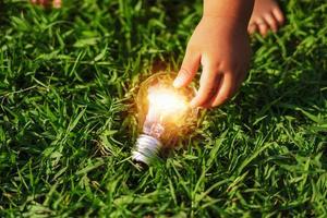 mão de crianças segurando a lâmpada na grama verde. conceito eco energia foto