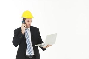 engenheiro bonito de terno e camisa branca e vestindo um chapéu amarelo de engenharia de segurança com a mão segurando laptop e smartphone falando e trabalhando em projeto isolado no fundo branco. foto