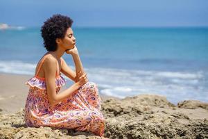 jovem negra sentada nas rochas na praia, olhos fechados, relaxando com técnicas de atenção plena. foto