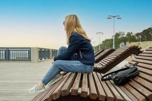 uma jovem loira solitária em um aterro vazio da cidade repousa sentado em uma espreguiçadeira em uma noite de primavera ou outono e olha para longe. a menina sente falta do calor e do verão. foto