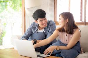 jovem casal asiático usando laptop pensa e pesquisando na internet juntos, homem e mulher casual sorrindo trabalham em casa com feliz e relaxe, comunicação e conceito de negócios. foto