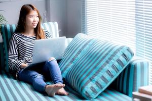 bela do retrato jovem mulher asiática usando laptop para lazer no sofá na sala de estar, garota trabalhando online com notebook freelance com um conceito de negócio de comunicação feliz. foto