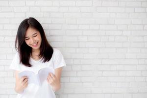 linda de retrato jovem mulher asiática felicidade relaxe em pé lendo livro sobre fundo branco de cimento concreto em casa, menina feliz estudo conteúdo literatura, educação e conceito de estilo de vida. foto
