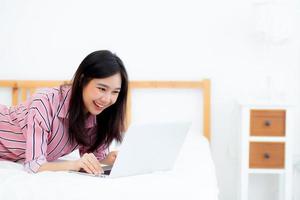 bela jovem asiática deitada na cama usando laptop no quarto para lazer e relaxar, freelance com notebook de trabalho de garota, conceito de comunicação. foto