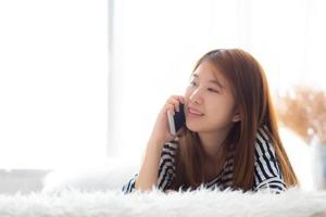 bela jovem asiática sorrindo deitado relaxe na cama de manhã, garota usando telefone inteligente móvel falando desfrutar, comunicação e conceito de rede social. foto