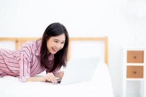 bela jovem asiática deitada na cama usando laptop no quarto para lazer e relaxar, freelance com notebook de trabalho de garota, conceito de comunicação. foto