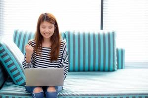 linda de retrato jovem asiático animado e feliz com o sucesso com laptop no sofá na sala de estar, menina trabalhando na sala de estar em casa, conceito de negócio freelance de carreira. foto