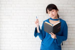 linda mulher asiática sorrindo em pé pensando e escrevendo caderno sobre fundo branco de cimento concreto em casa, lição de casa de menina no conceito de livro, educação e estilo de vida. foto