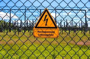 sinal mostrando as palavras de alta tensão em língua alemã na cerca de uma grande subestação de distribuição de energia elétrica com muitas linhas de energia em um dia ensolarado foto