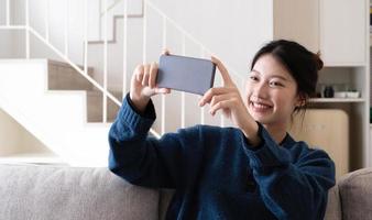 foto de uma jovem feliz da Ásia tomando selfie com seu celular enquanto está sentado na sala de estar.