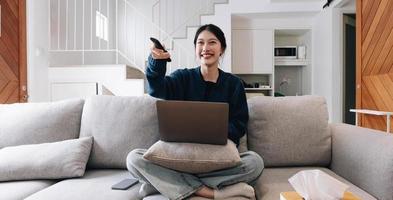 bela jovem asiática assistindo filme divertido na televisão em casa. garota casual atraente sinta-se feliz e relaxe, sente-se no sofá se divertindo assistindo vídeo de comédia na tv em casa. conceito de estilos de vida de atividade foto