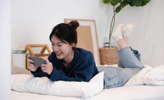 retrato bela jovem mulher asiática em um colchão branco no quarto de manhã. o smartphone está nas mãos das mulheres. senhora segurando o telefone.