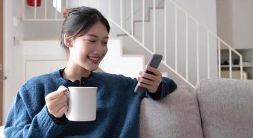 retrato de uma mulher asiática sorridente sentada no sofá e tomando café olhando para smartphone. foto