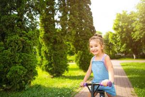 uma garota de macacão jeans anda de bicicleta roxa no verão em um parque verde. entretenimento ativo para crianças, scooter para crianças, bebê feliz. espaço de cópia foto