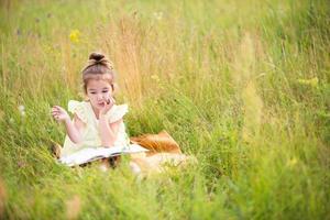 garota de vestido amarelo senta-se na grama em um cobertor em um campo e lê um livro de papel. dia internacional da criança. horário de verão, infância, educação e entretenimento, núcleo de chalé. espaço de cópia foto