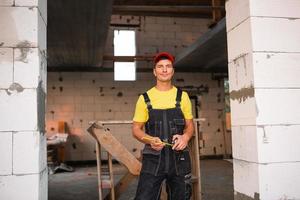 engenheiro trabalhador da construção civil no canteiro de obras mede o comprimento da abertura da janela e parede de tijolos com fita métrica. homem em roupas de trabalho - macacão e boné foto