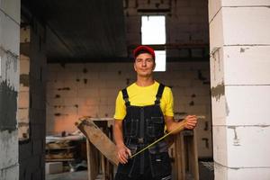 engenheiro trabalhador da construção civil no canteiro de obras mede o comprimento da abertura da janela e parede de tijolos com fita métrica. homem em roupas de trabalho - macacão e boné foto