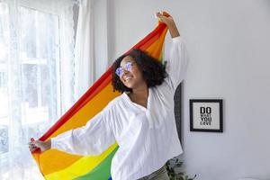 garota afro-americana segurando a bandeira do arco-íris lgbtq em seu quarto para sair do armário e celebração do mês do orgulho para promover a diversidade sexual e igualdade no conceito de orientação homossexual foto