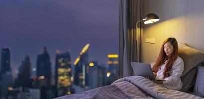 mulher asiática trabalhando on-line com laptop em sua cama tarde da noite com a vista da cidade do edifício de arranha-céus para estilo de vida de luxo urbano e conceito de educação