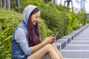 jovem mulher asiática relaxadamente sentada no parque público usando telefone celular para mídias sociais e bate-papo foto