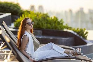 bem-sucedida jovem empresária asiática sentada na cama da piscina olhando a vista durante o pôr do sol para férias de verão elegantes e conceito de estilo de vida de luxo urbano com vista para a paisagem urbana