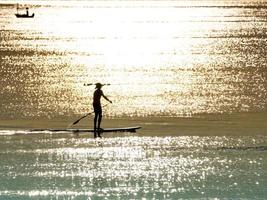 homem de silhueta em pé no paddleboard no mar calmo com reflexo da luz do sol da manhã na superfície da água foto