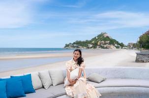 turista de mulher asiática feliz sente-se e segurando o travesseiro no sofá com vista para a praia do mar azul e montanha em huahin, tailândia, no verão foto