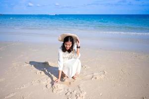 mulher asiática atraente de vestido branco gosta de sentar e jogar areia na bela praia da ilha foto