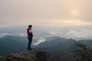 homem asiático com máscara facial protetora em pé na borda da rocha do pico da montanha no ponto de vista pha tang 104 com vista para o rio e névoa do mar e nascer do sol no céu de neblina pesada. viajar em chaing rai, tailândia foto