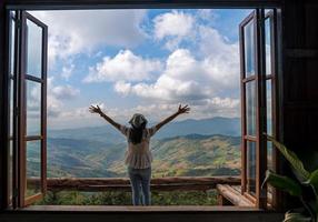parte traseira da mulher asiática com os braços da liberdade abertos em pé atrás da grande janela de madeira com vista para a montanha e céu azul nublado em doi chang, atração de viagens em chiang rai foto