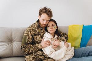 casal ucraniano, militar de uniforme com a namorada no sofá em casa em um fundo de bandeira amarela e azul foto