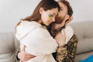 casal ucraniano, militar de uniforme beija sua namorada no sofá em casa em um fundo de bandeira amarela e azul foto