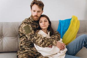 casal ucraniano, militar de uniforme com a namorada no sofá em casa em um fundo de bandeira amarela e azul foto