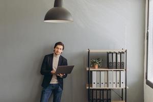 imagem do jovem empresário alegre de terno segurando e usando laptop no escritório moderno foto